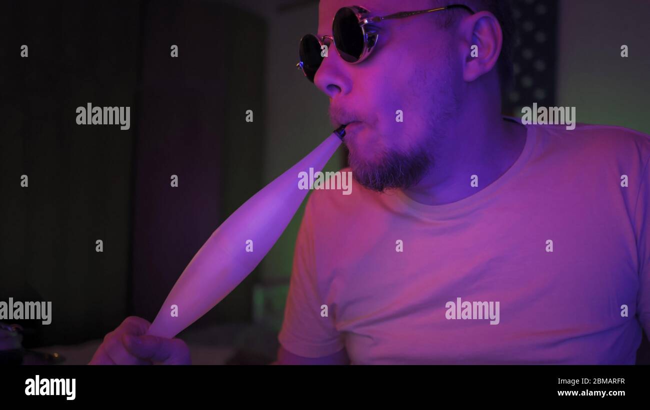 Man inhales the smoke of a hookah In Neon Lighting, Close Up Portrait of caucasian smoker in in dark room with fluorescent glowing. Quarantine at home Stock Photo