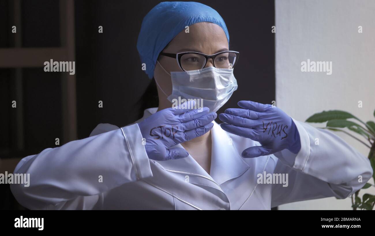 Tired female doctor in white uniform and protective mask shows the inscription stop virus on his palms in rubber gloves looking to the side Stock Photo