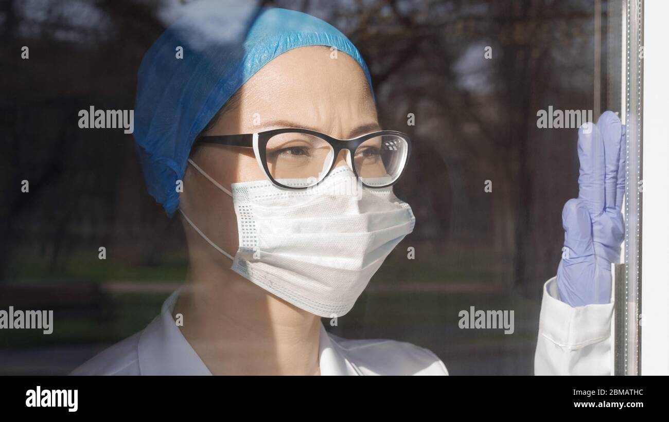 Tired Doctor looks at window Touch Glass With Hand. Female medic thoughtfully awaiting the end of the pandemic. Medicine and healthcare concept Stock Photo