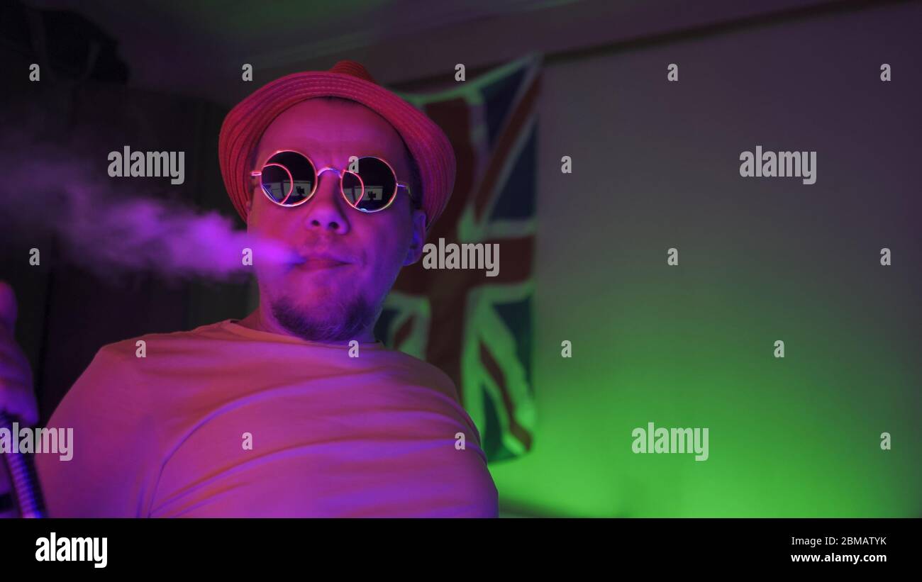 Smoker of Hookah Exhaling Smoke Up In Neon Lighting. Man in sunglasses in self-isolation at home smokes in room with neon lights on GB flag Background Stock Photo