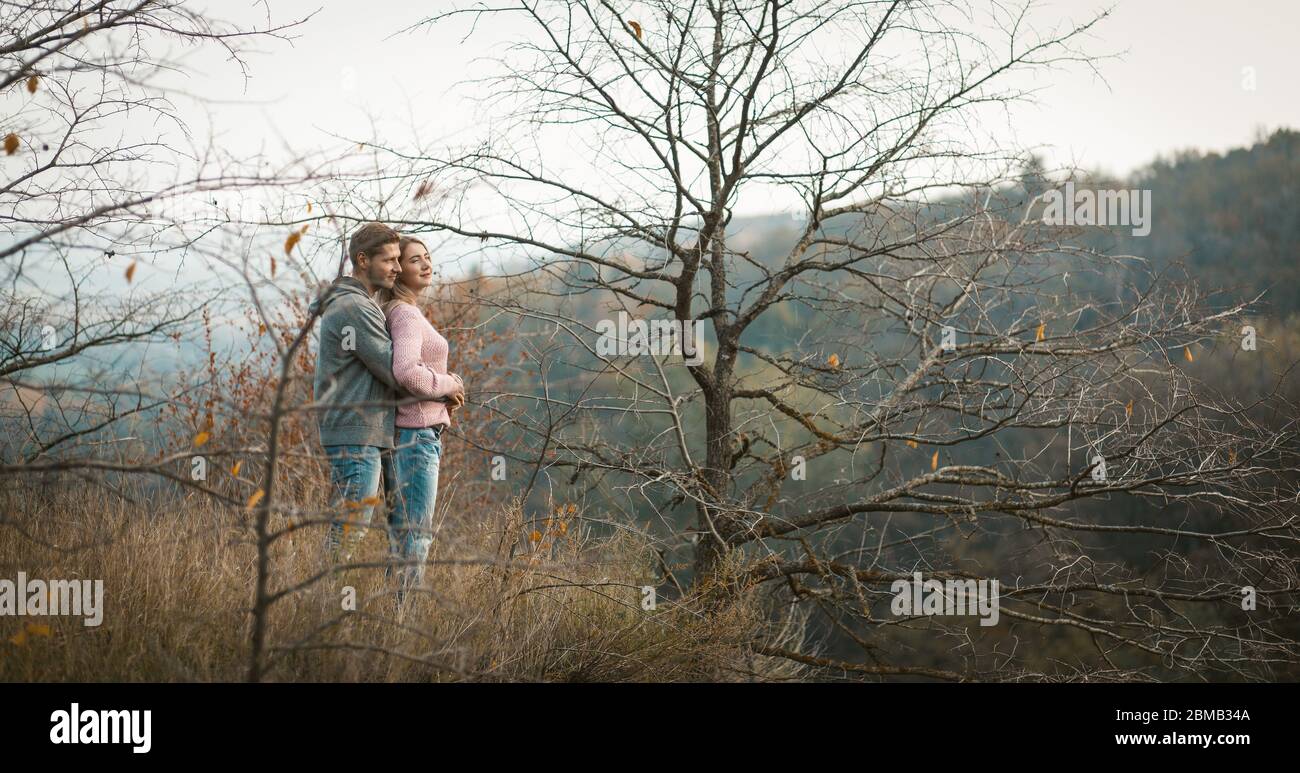Hugging couple in love stands on a hillside looking down, young man and a woman admire the autumn forest growing on the slopes below Stock Photo