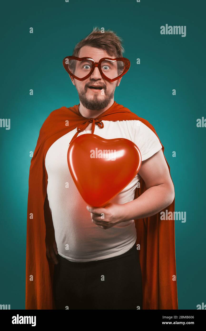 Superman In Love Holds Big Red Heart. Positive Man In Superhero Costume And Heart-Shaped Glasses Poses On Biscay Green Background. Valentine's day Stock Photo