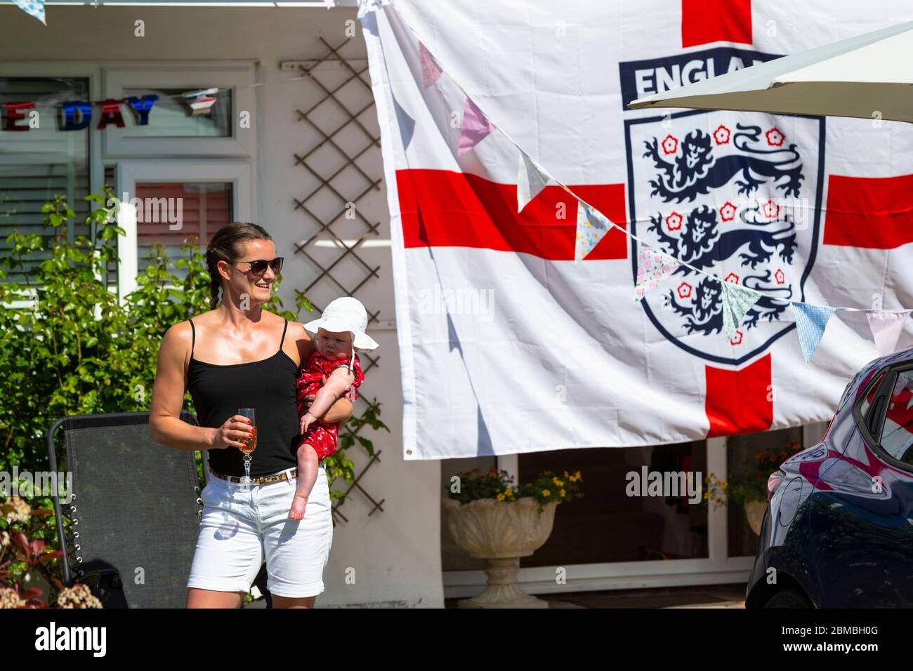 Ashford, Kent, UK. 8th May, 2020. Members of the community in the village of Hamstreet near Ashford in Kent come together to commemorate VE day on it's 75th anniversary with a tea party outside their front garden. A woman drinking champagne holding a baby beside a a large England flag. ©Paul Lawrenson 2020, Photo Credit: Paul Lawrenson/Alamy Live News Stock Photo