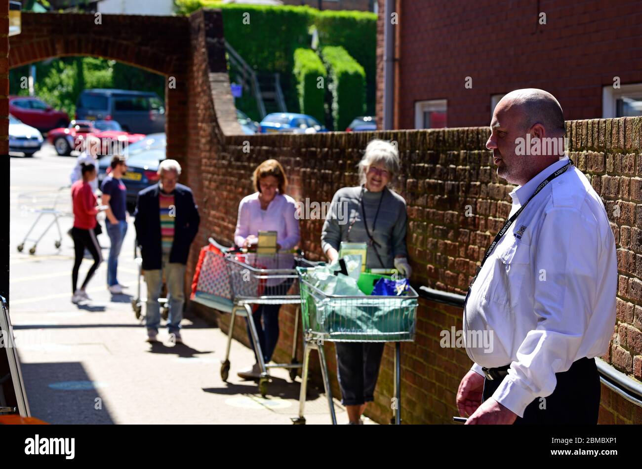 Security guard (foreground right) ensuring shoppers enter supermarket according to social distancing measures introduced due to the COVID-19, Surrey Stock Photo