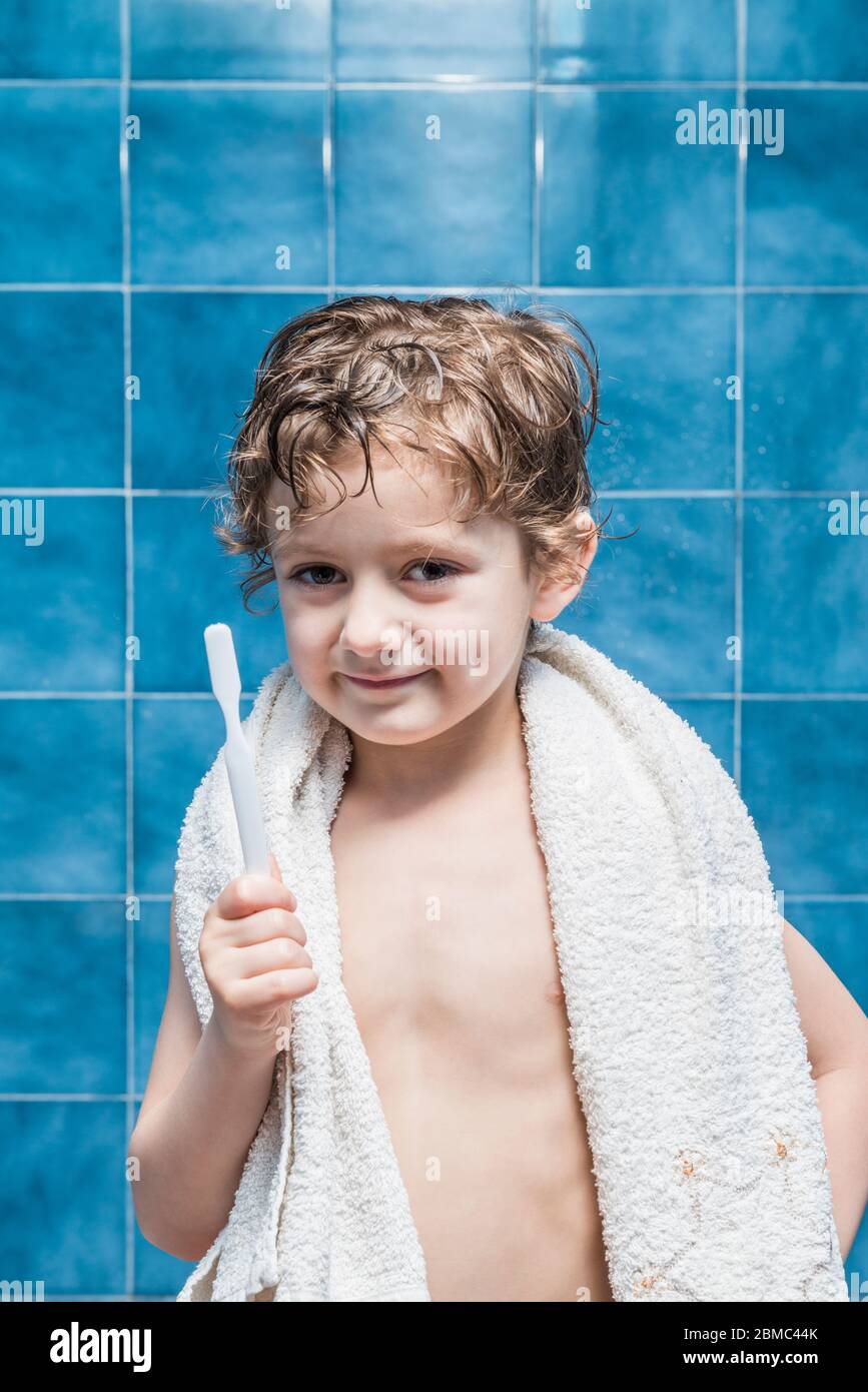 A child with a towel on his shoulders showing a toothbrush, with blue tiles in the background. Stock Photo