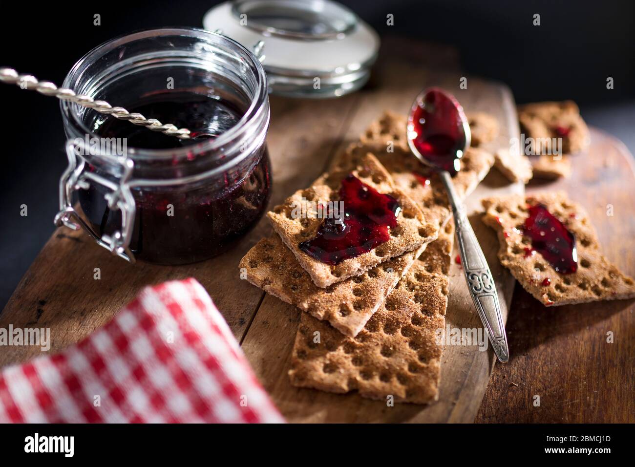 Open jar with blackcurrant jam on a wooden cutting board. Dry biscuits spread with jam, crumbs and a spoon in jam. Space for text. Stock Photo
