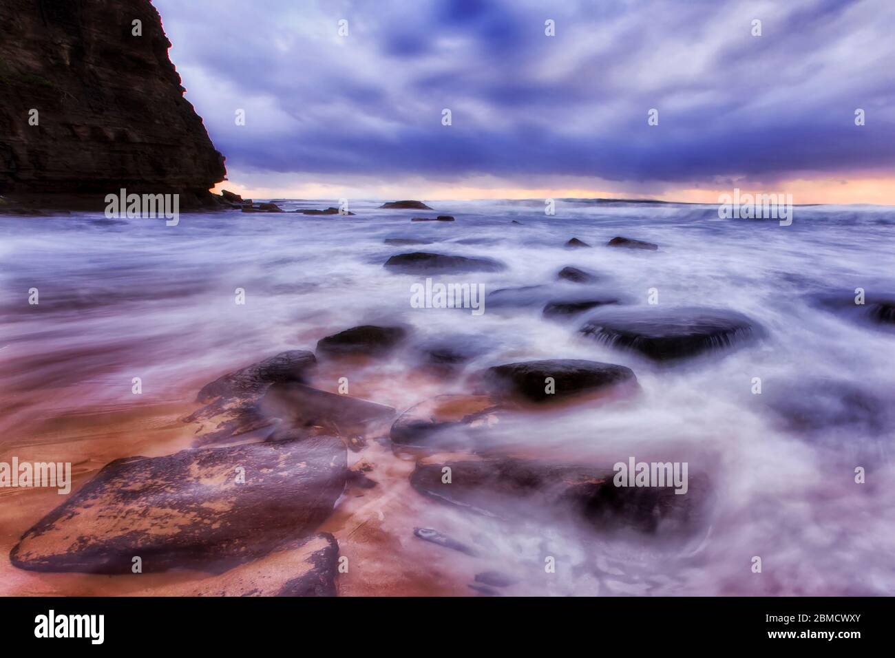 ROcks on sandy beach of Sydney Northern beaches near tall headland cliff at sunrise during stormy weather. Stock Photo