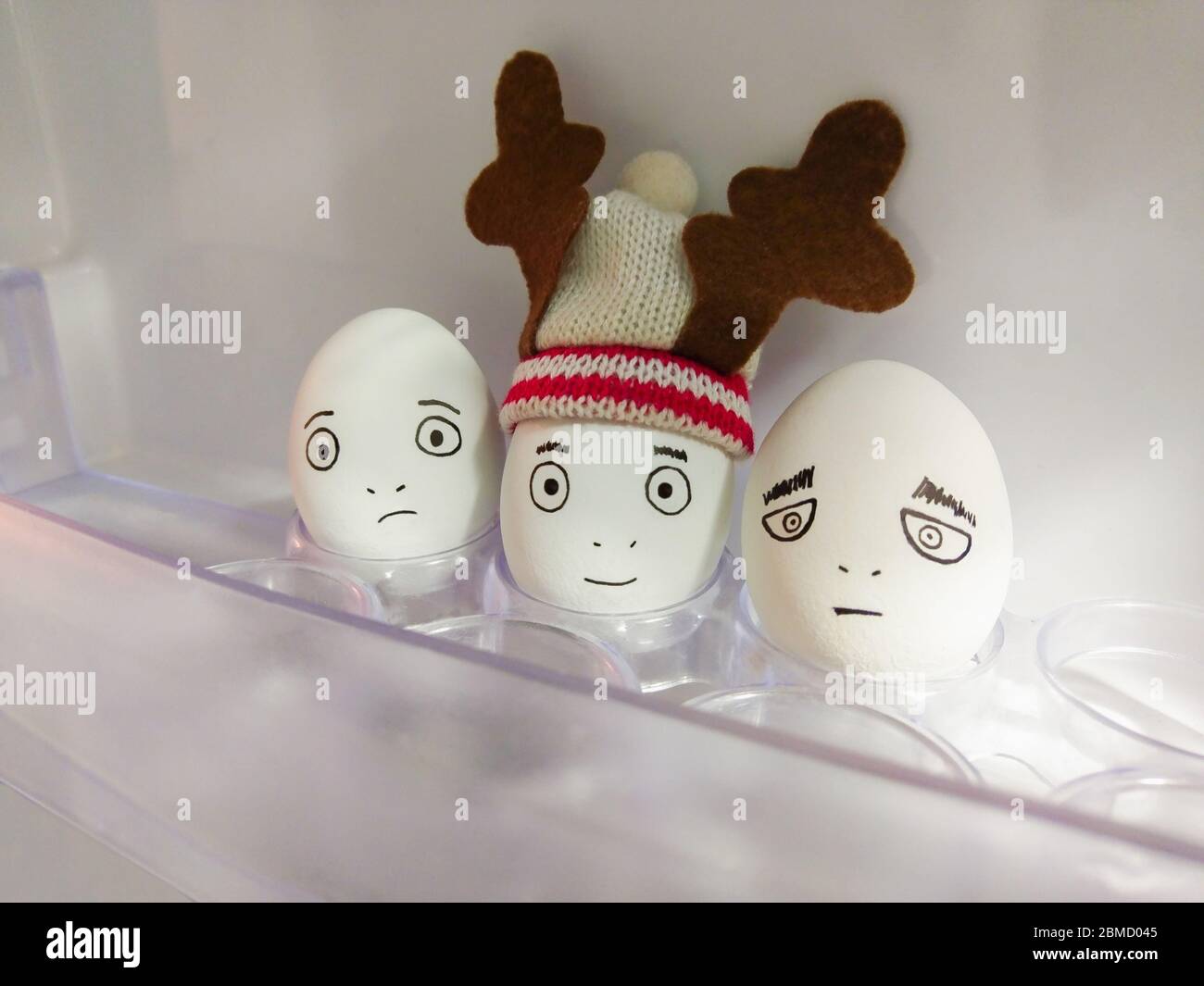 Three white chicken eggs with faces drawn on them. Central egg wearing funny warm hat with plush antlers. Fridge door interior. Funny decorating idea. Stock Photo
