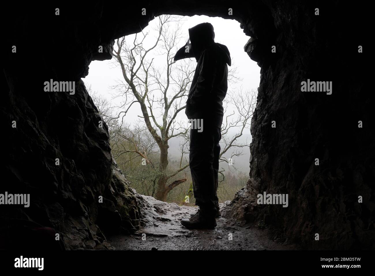 A scary, hooded figure wearing a plague doctor mask, standing in the entrance of a cave on a misty winters day Stock Photo