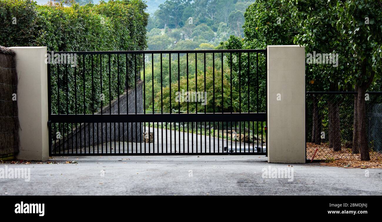 Black metal wrought iron driveway property entrance gates set in brick fence, concrete path, garden trees Stock Photo
