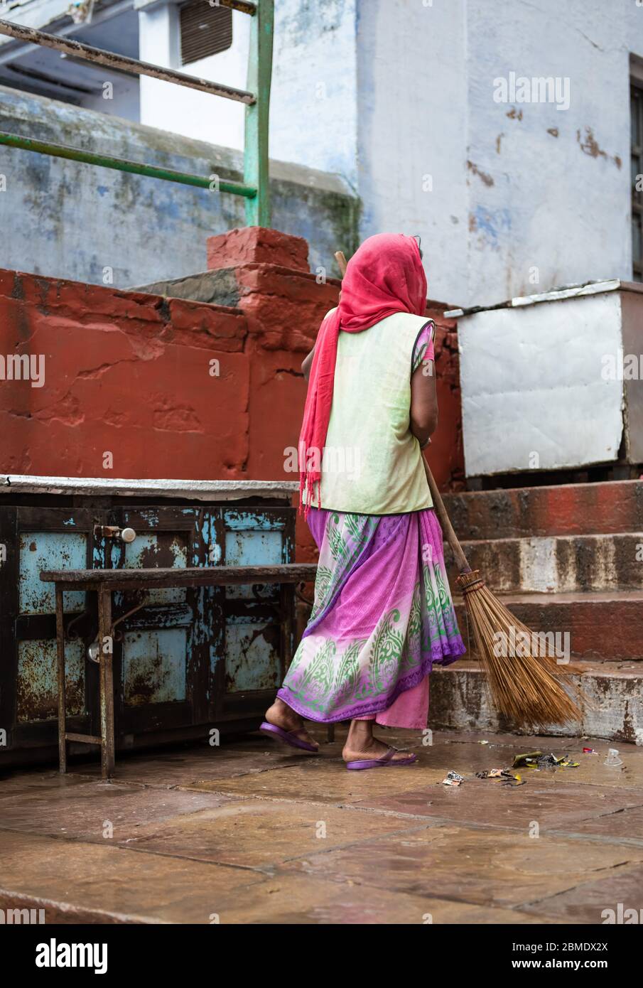 One woman sweeping a dirty walkway in Varanasi, India, wearing a head covering and colorful clothing, near the Ganges River. Stock Photo