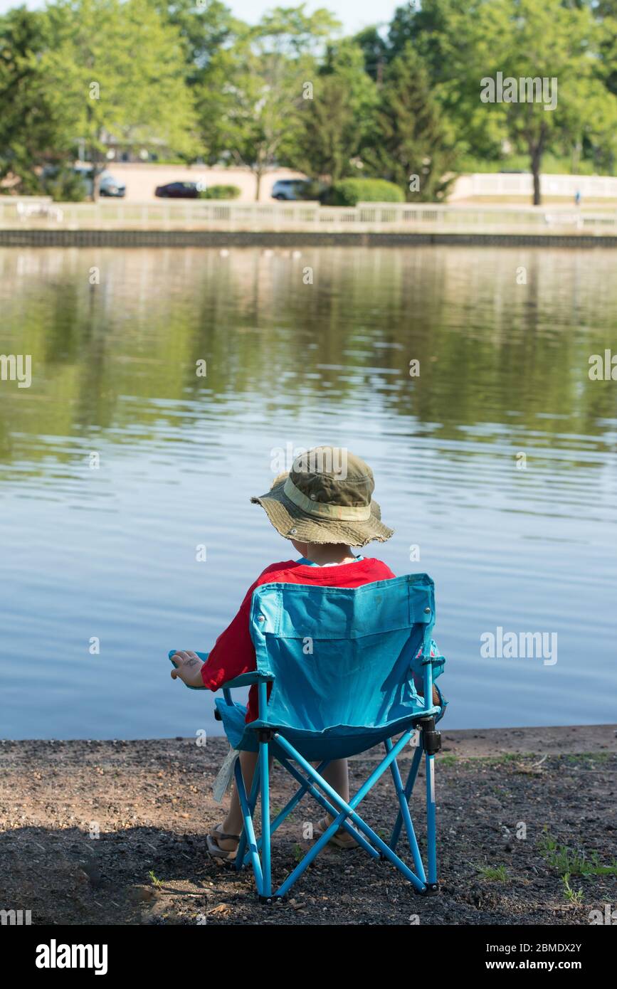 Young, caucasian boy wearing a fishing hat, sitting in a little, blue chair looking out at the water on a lake in a park. Stock Photo