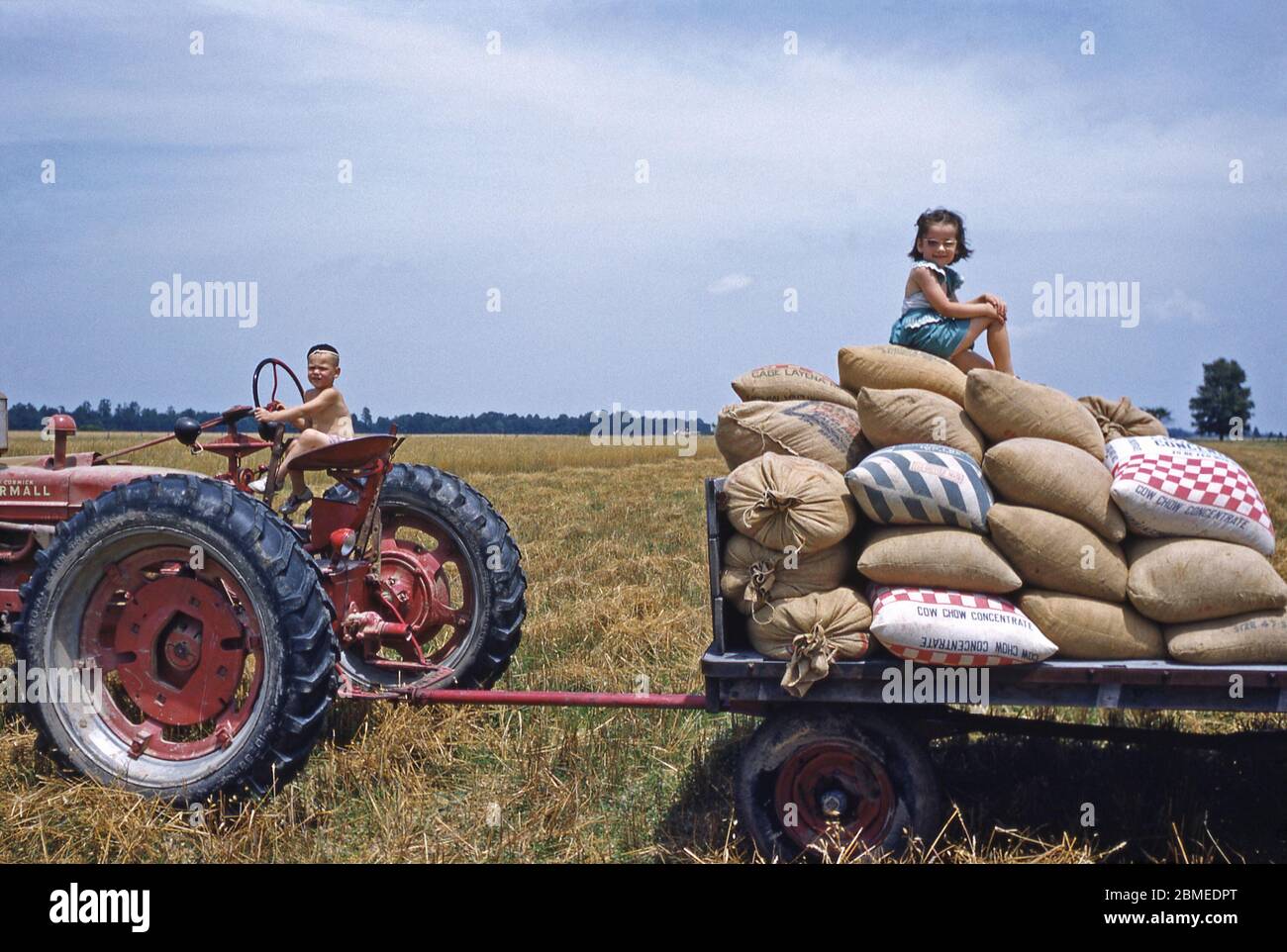 Two children playing in the sunshine on a tractor and trailer on the family farm, USA in the early 1950s. The red and blue sacks on the trailer contain 'Chow Chow Concentrate', an animal feed supplement. Stock Photo