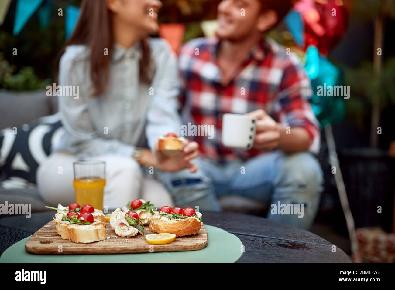 focus on sandwiches with cherry tomato and orange juice with couple in the background out oif focus talking, smiling. People, food and beverage Stock Photo