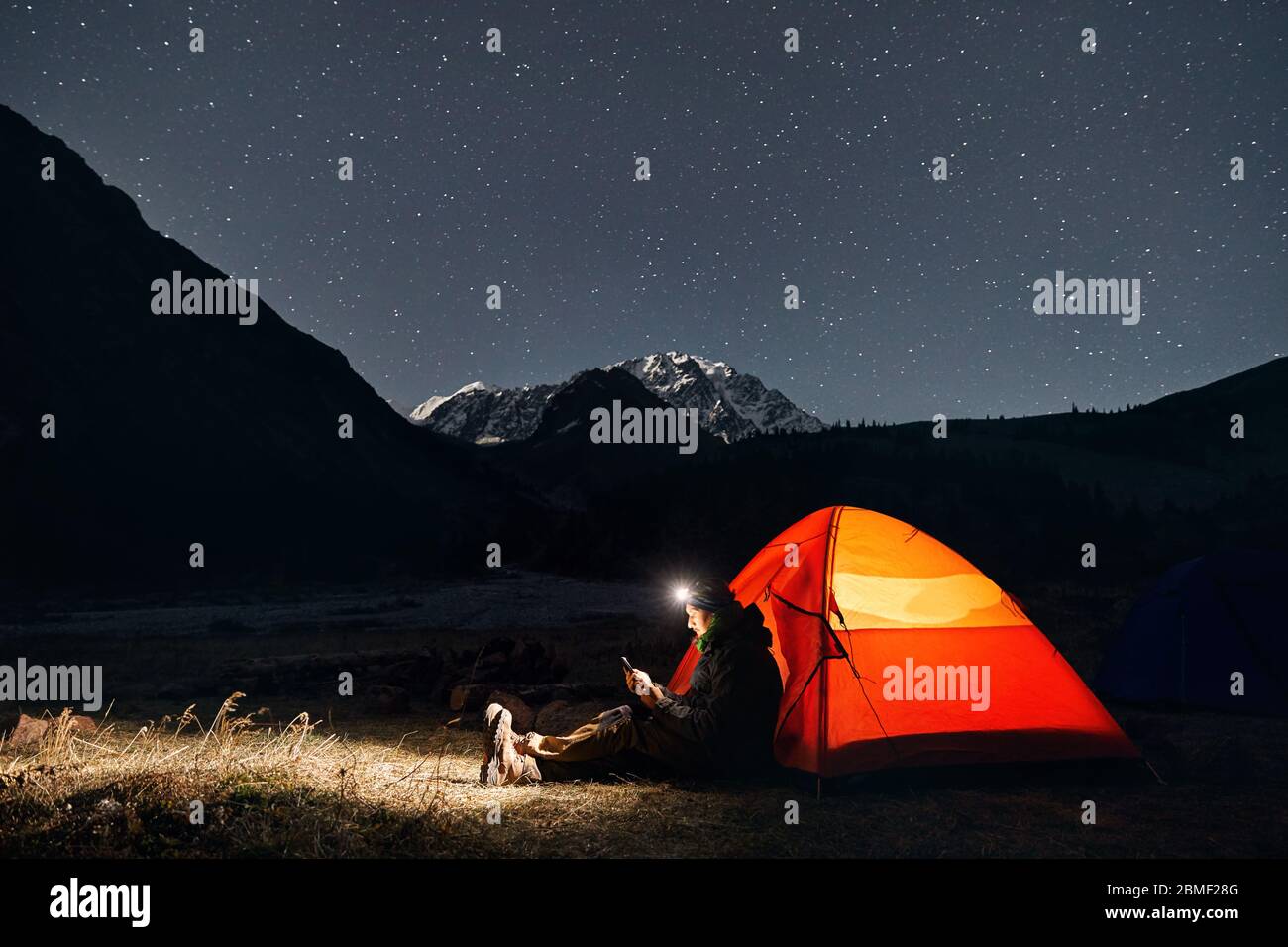 Man with headlamp near glowing orange tent in the mountains under night sky with stars Stock Photo
