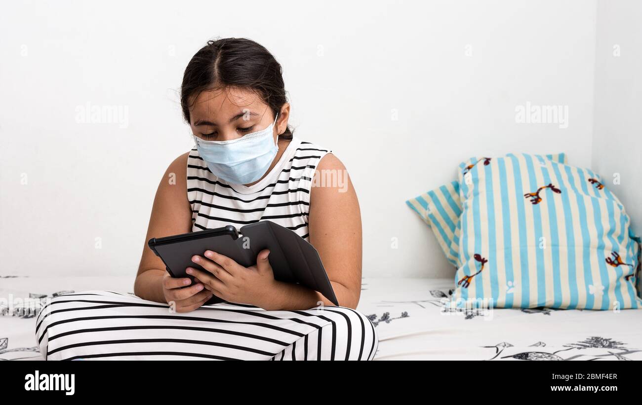 Young Asian girl wearing protective face mask sitting on her bed using a tablet to study online - corona virus home schooling concept image with copy Stock Photo