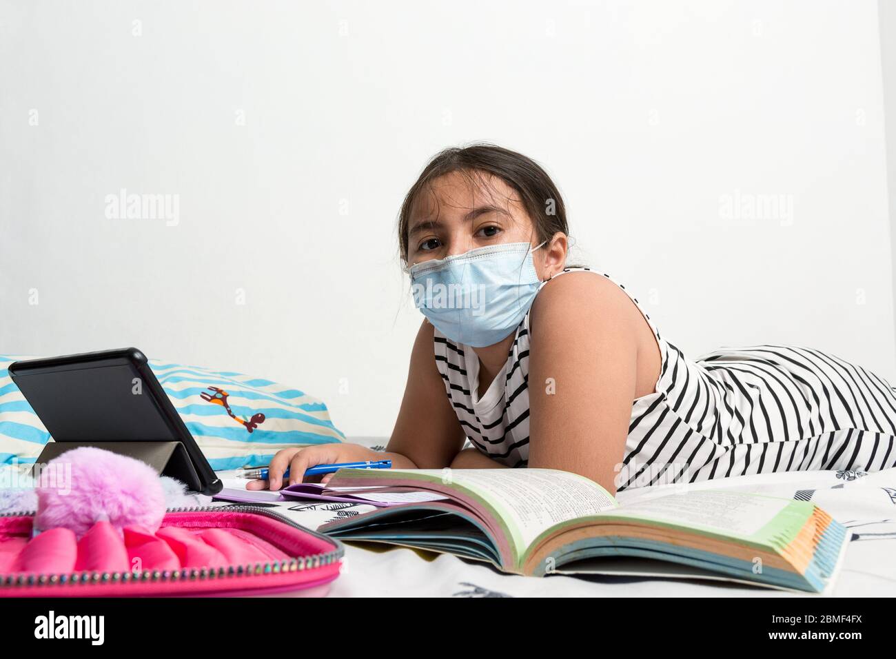 Young Asian girl wearing protective face mask laying on her bed studying - corona virus home schooling concept image with copy space for text Stock Photo