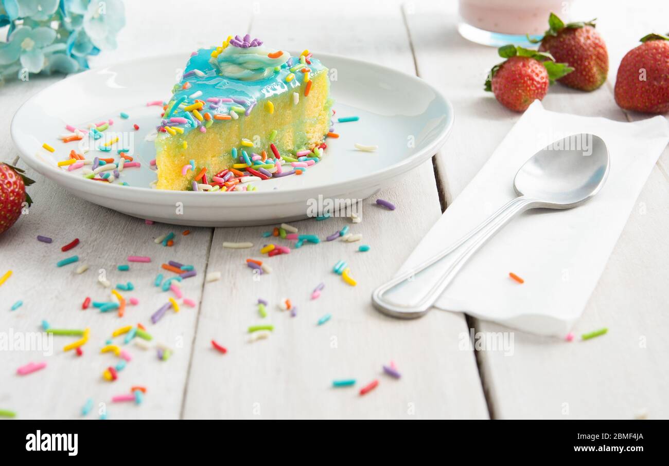 A slice of colorful sponge cake with confetti sprinkles a spoon and some strawberries -  kitchen tabletop food, baking and lifestyle concept image. Stock Photo