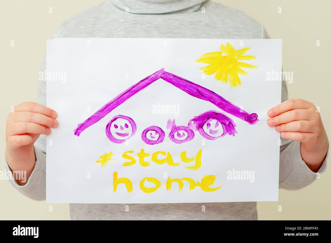 Hands of child holding a picture of family silhouette under the roof and words Stay Home on yellow background. Children's creativity concept. Stock Photo
