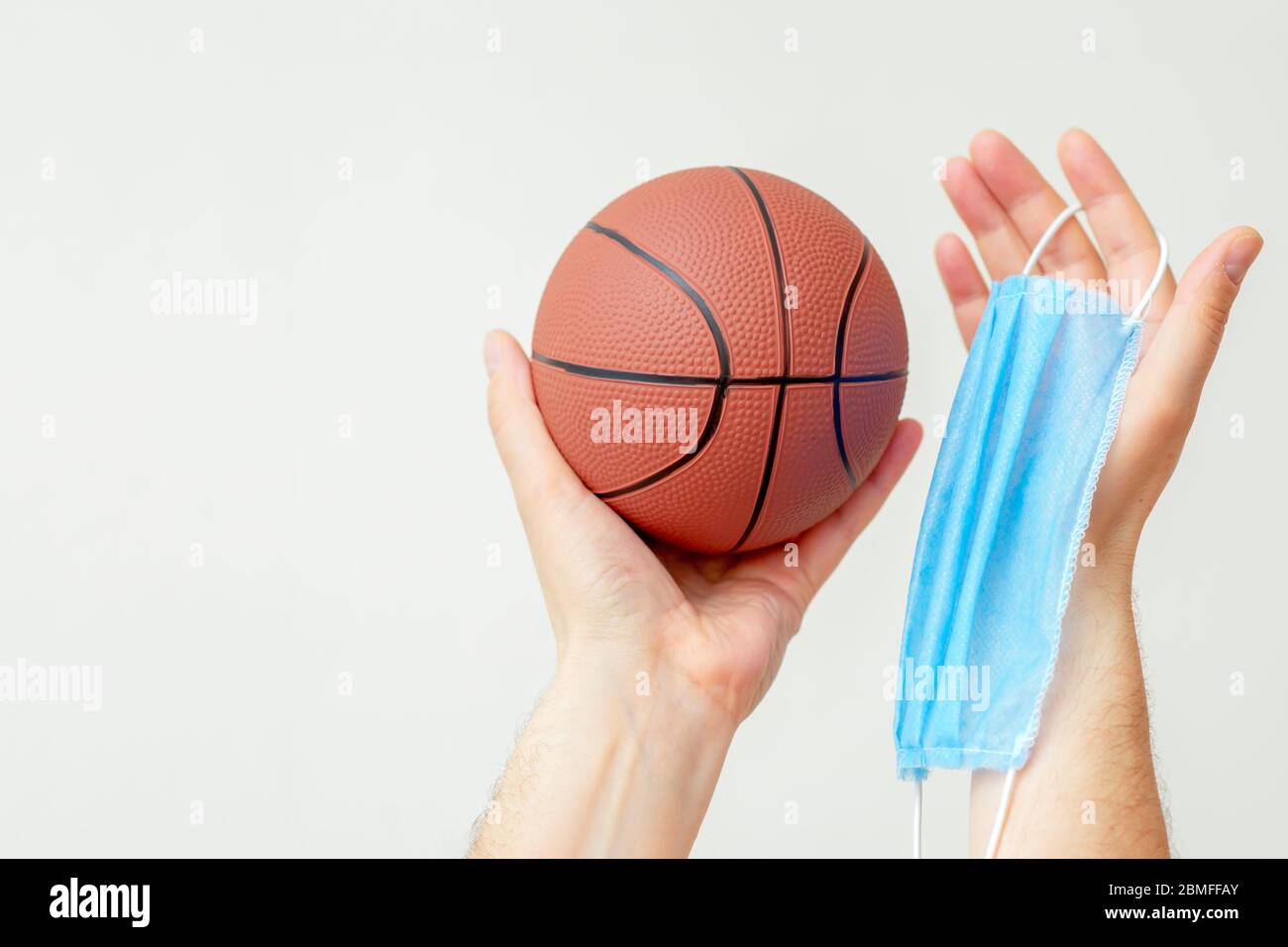 Hands of man holding basketball ball with medical mask on a light background with copy space. Coronavirus suspends world sport. Stock Photo