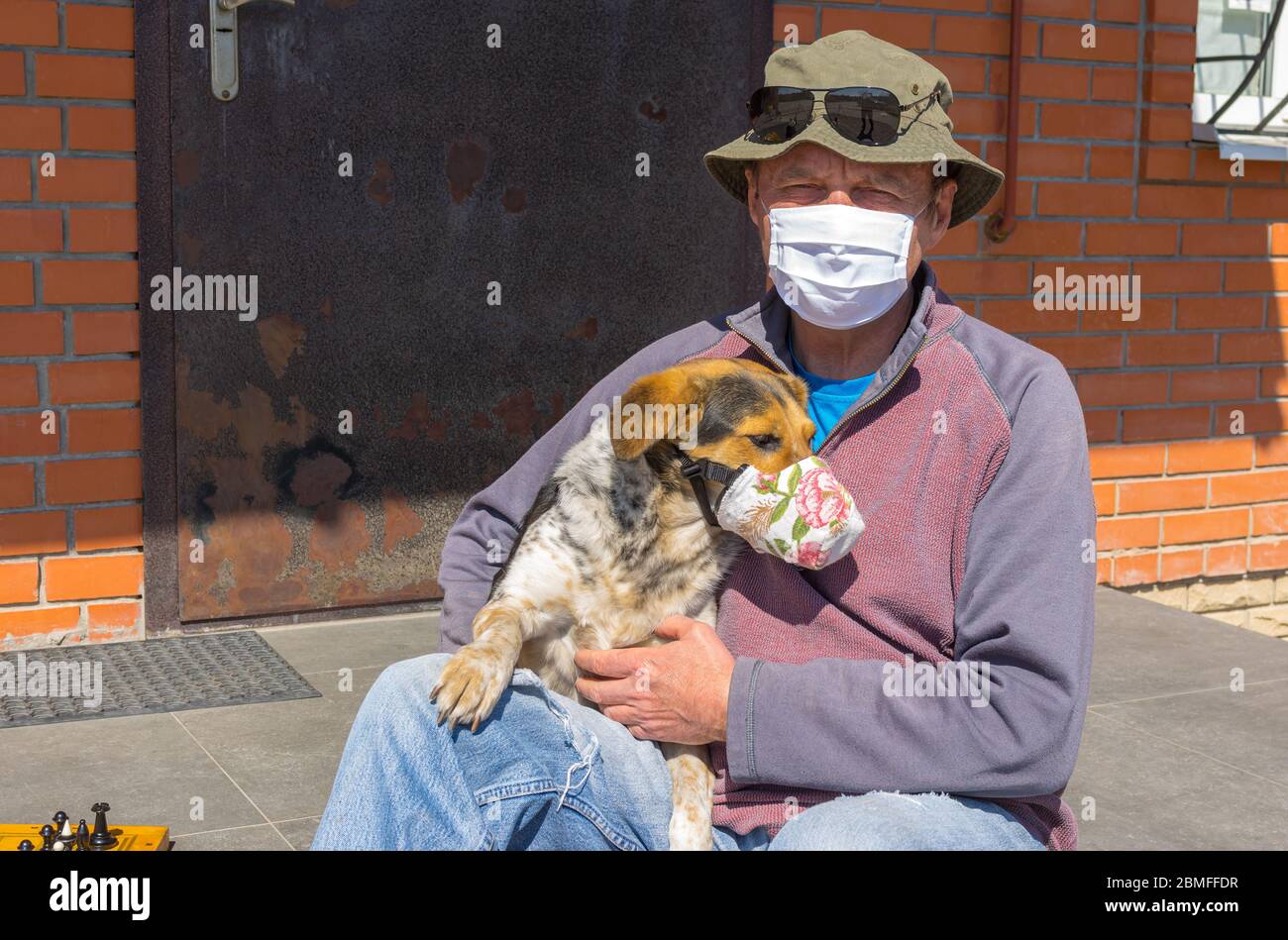 Portrait of Caucasian senior man with mixed breed dog wearing masks sitting on a  threshold while being on Coronavirus COVID-19 quarantine period in U Stock Photo