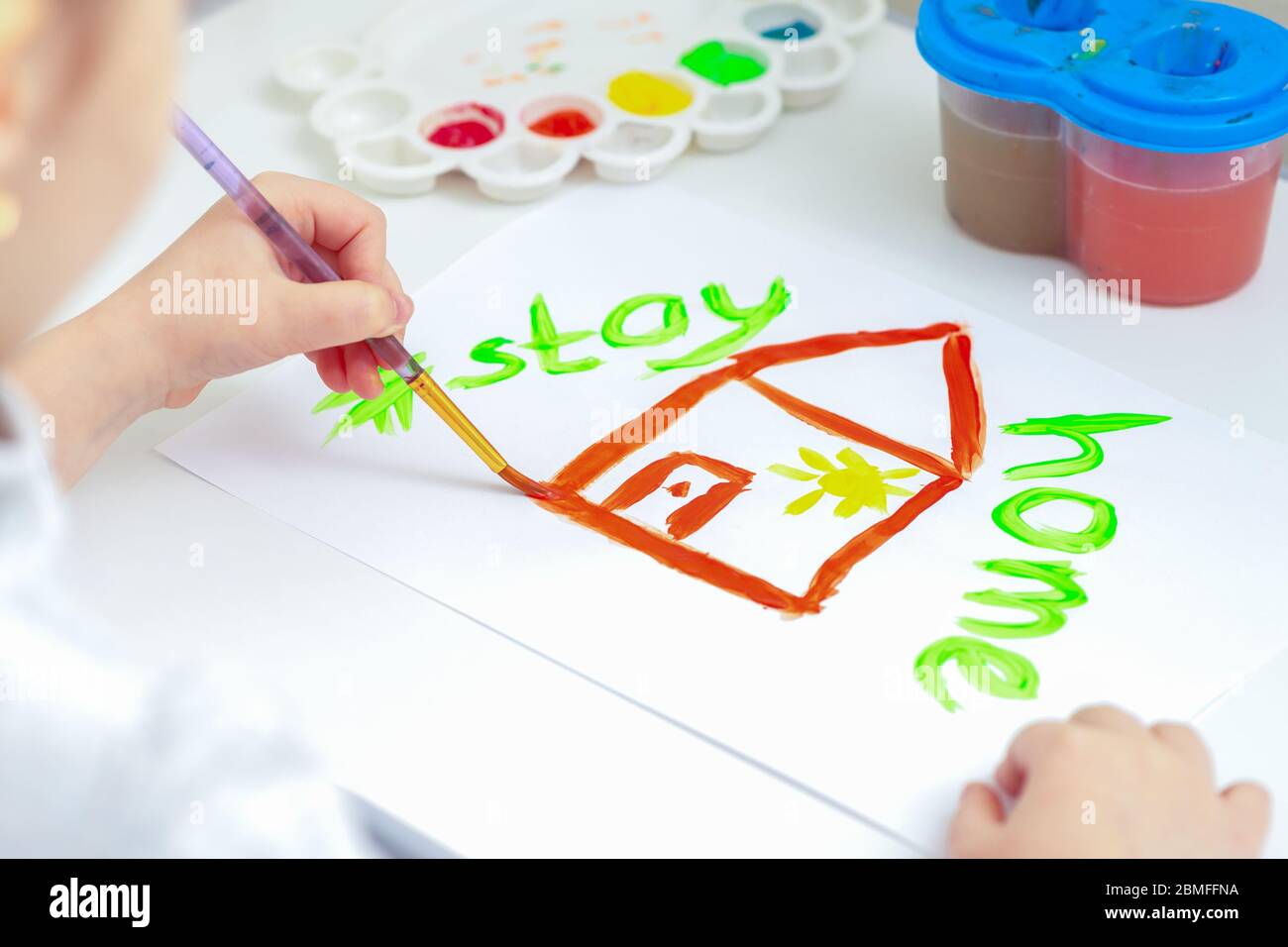 Child is drawing house and Stay Home words by brush with watercolors on white paper at home. Stock Photo