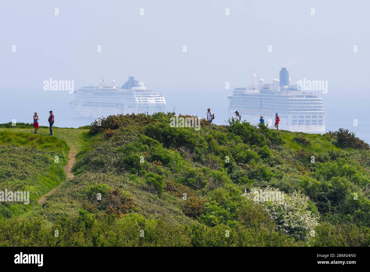 Weymouth, Dorset, UK. 9th May 2020. UK Weather: Walkers on the cliff top on the South West Coast Path with a view of the five cruise ships anchored in the bay off the coast of the seaside resort of Weymouth in Dorset on a hot sunny day due to the cruising industry shutting down during the coronavirus lockdown. There are four P&O ships which are The Oceana, Aurora, Arcadia and Ventura and one Cunard ship the liner Queen Mary 2. (Oceana and Arcadia pictured) Picture Credit: Graham Hunt/Alamy Live News Stock Photo