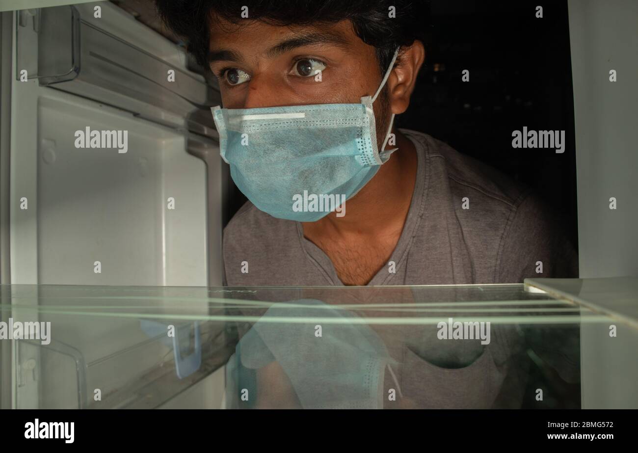Man in a medical mask looks into empty fridge or refrigerator for food - Concept of no pantry food available during home quarantine at covid-19 or Stock Photo
