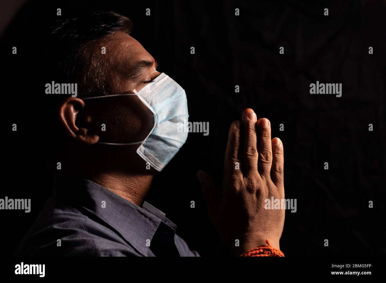 Man with medical mask praying to god by closing eyes in dark room to protect or save from covid-19 or coronavirus crisis - spirituality and Surrender Stock Photo