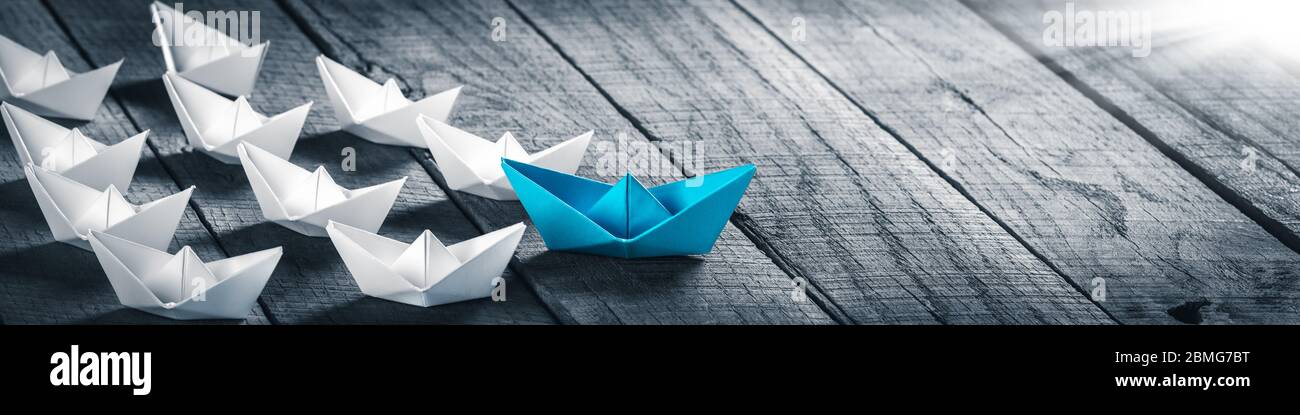 Blue Paper Boat Leading A Fleet Of Small White Boats On Wooden Table With Sunlight - Leadership Concept Stock Photo