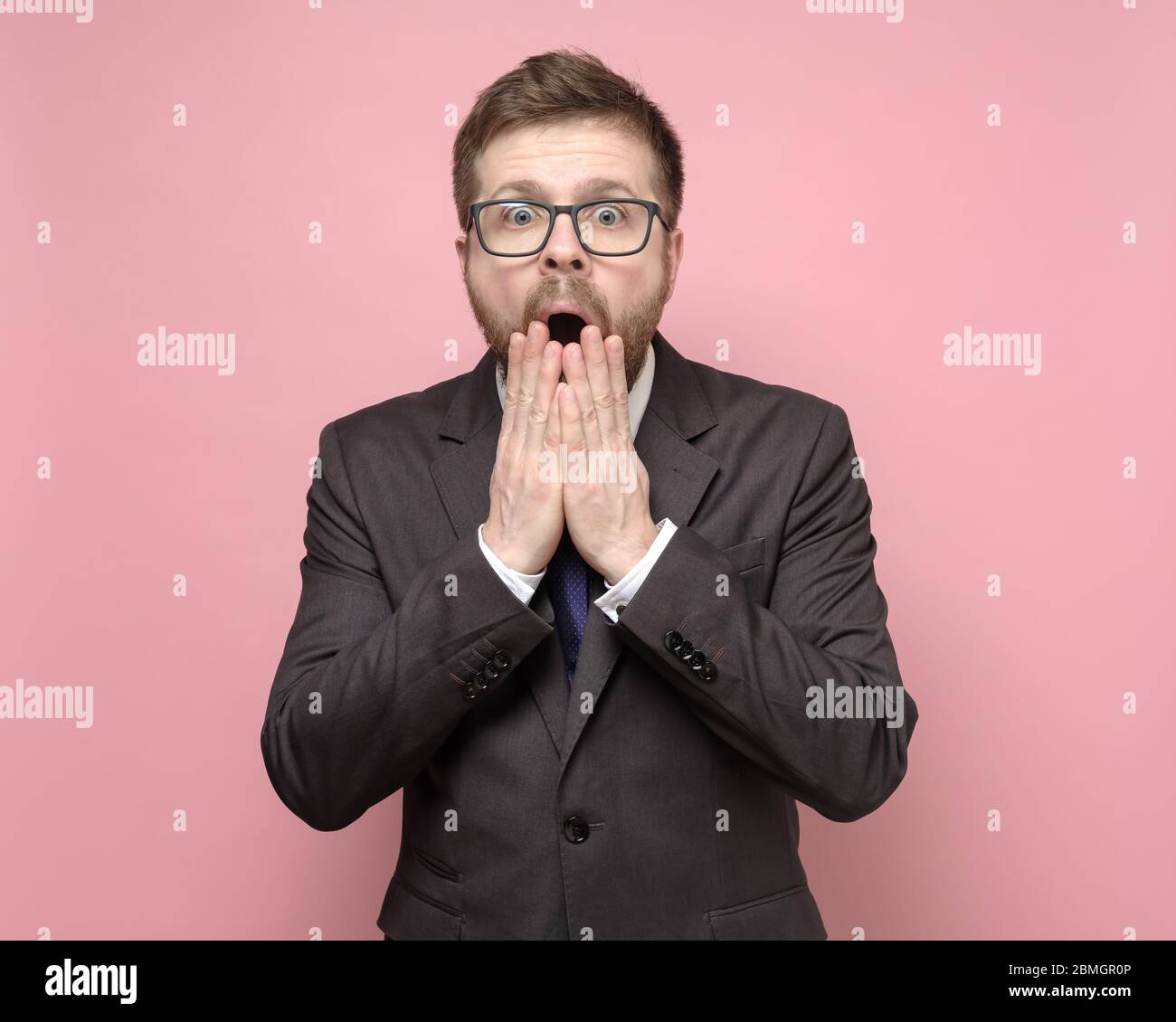 Surprised man with glasses and a suit looks at the camera with big eyes and covers his open mouth with hands. Stock Photo