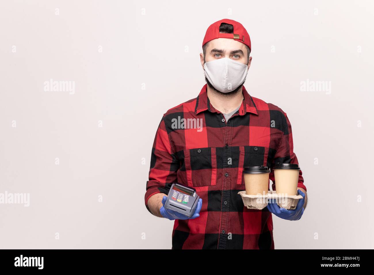 Young male waiter or courier of takeaway cafe in workwear, mask and gloves holding two glasses of coffee and payment machine in isolation Stock Photo