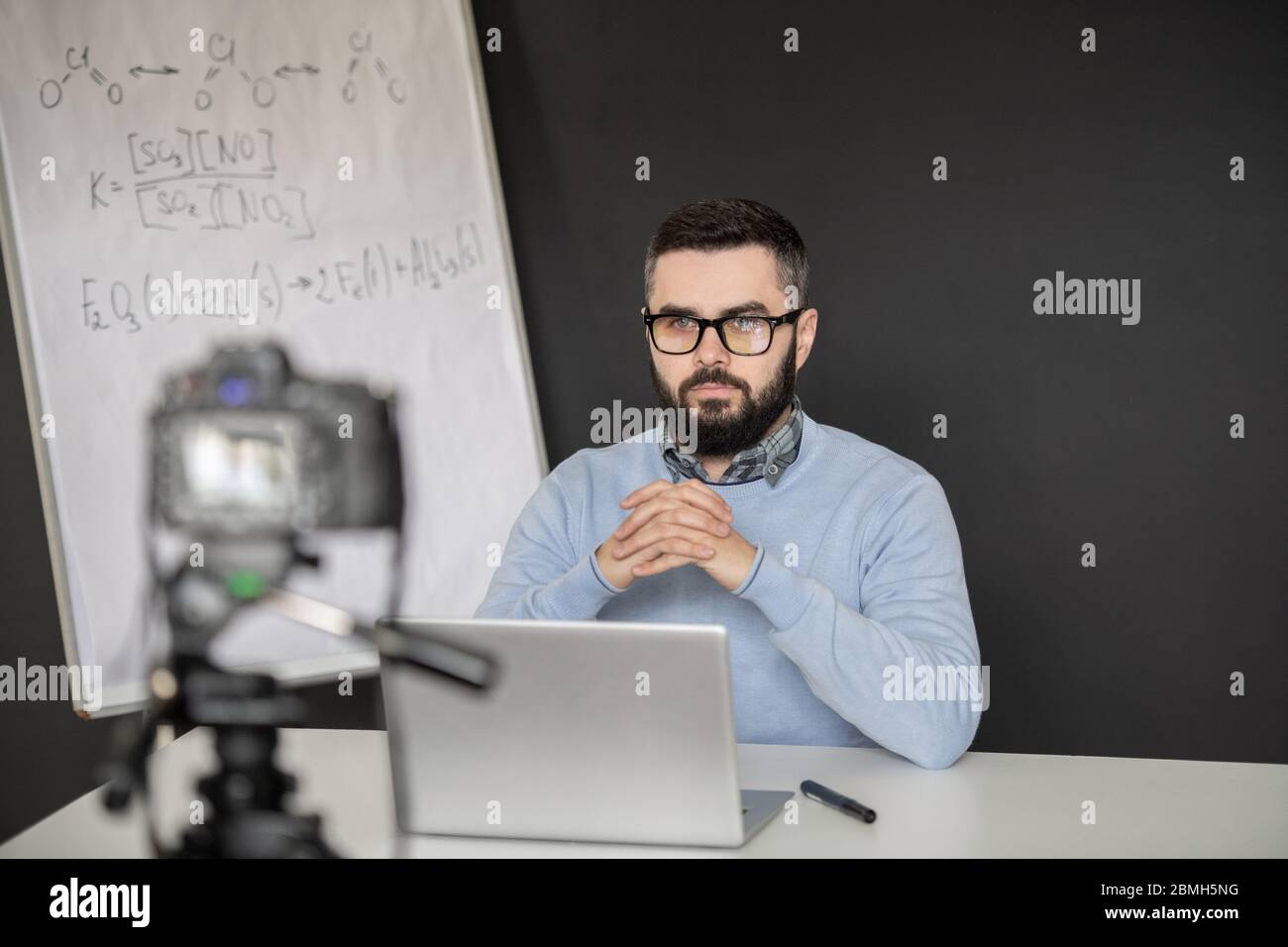 Serious bearded man holding hands by his face while looking at laptop display during remote lesson by whiteboard with chemical formulas Stock Photo