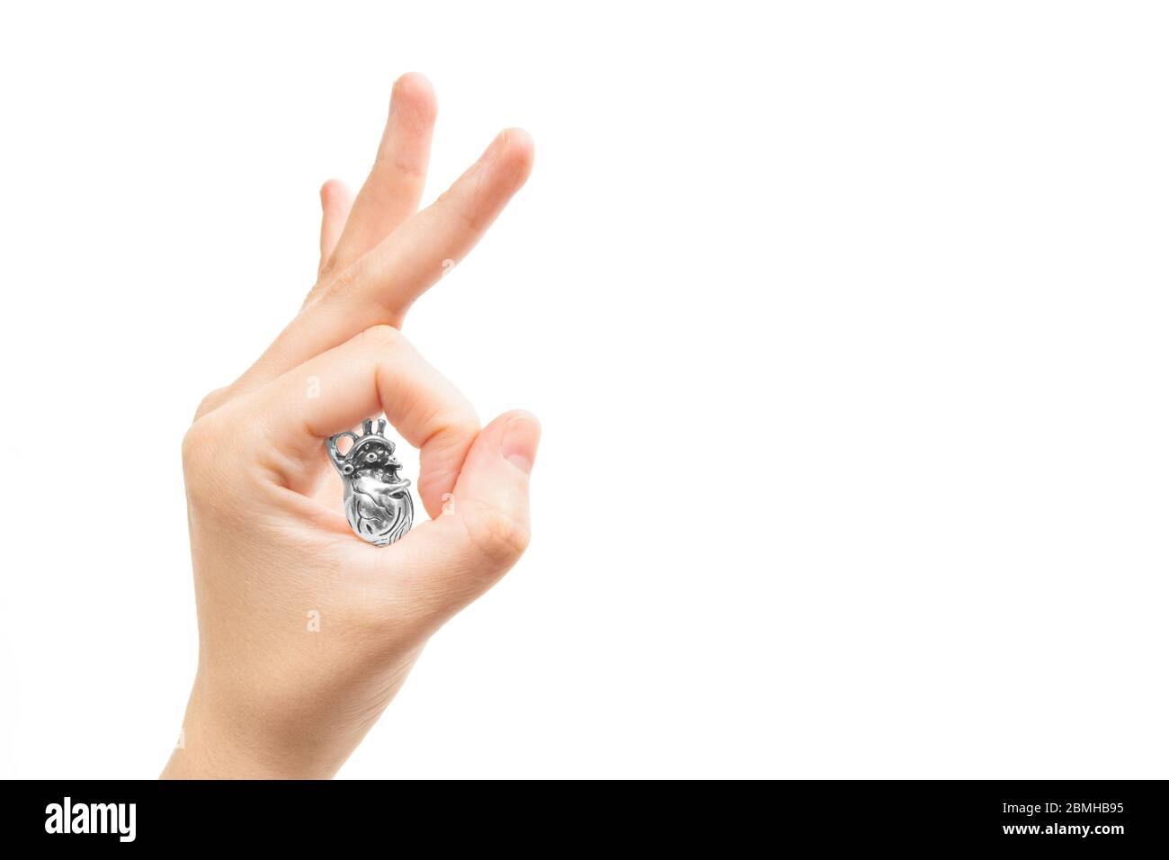 Female hand shows OK ring gesture holding a miniature steel copy of a human heart in the circle between the thumb and index fingers. Stock Photo