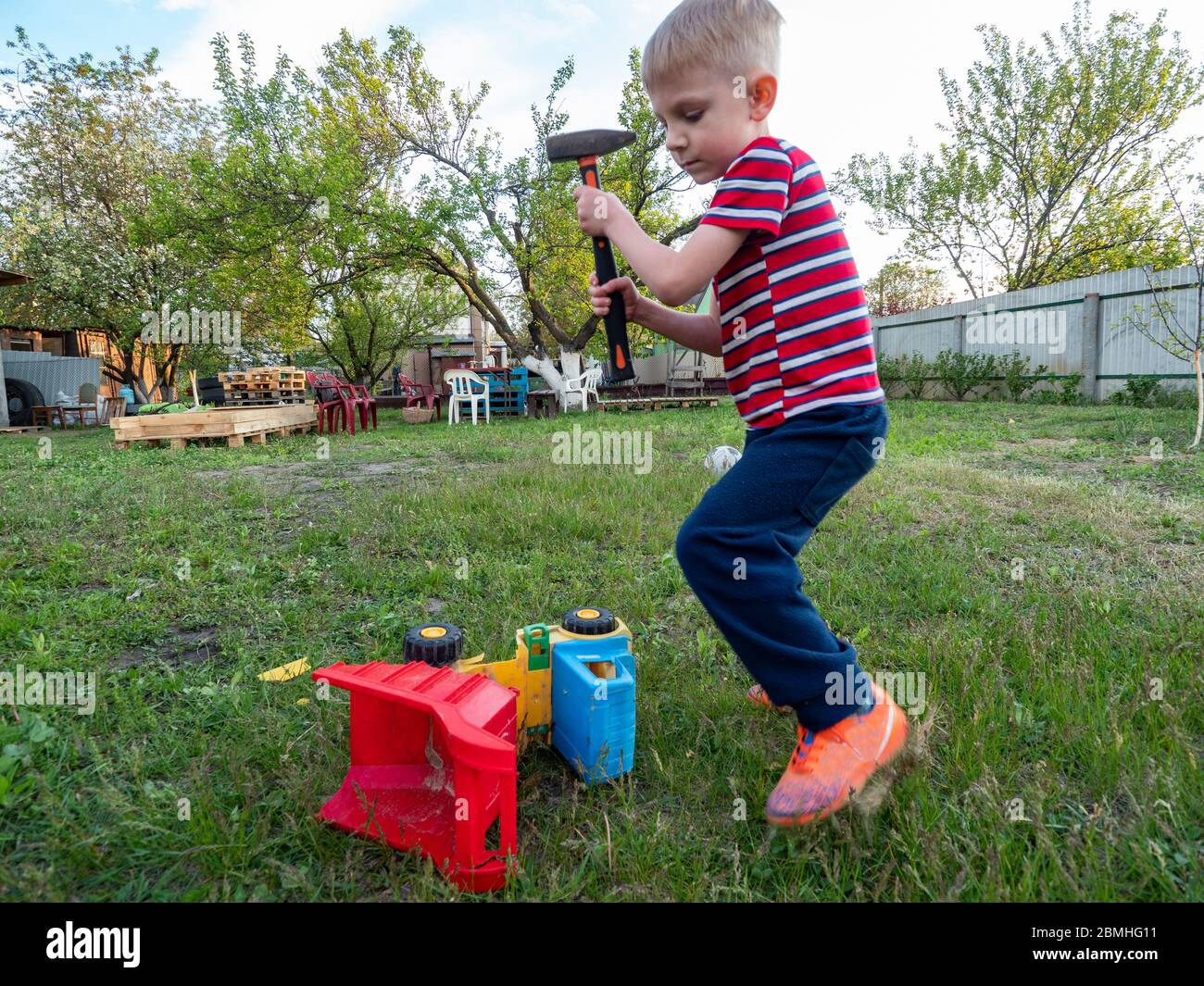 Little caucasian boy smashes a plastic toy car with a hammer in the garden Stock Photo