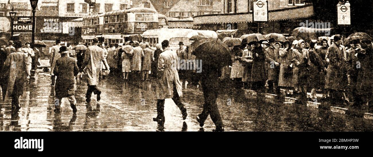 WEATHER - RAINY BRITAIN -Londoners in the rain. - 1950's street scene with passengers waiting in queues at a bus stop wearing headscarves, raincoats & carrying umbrellas. Stock Photo