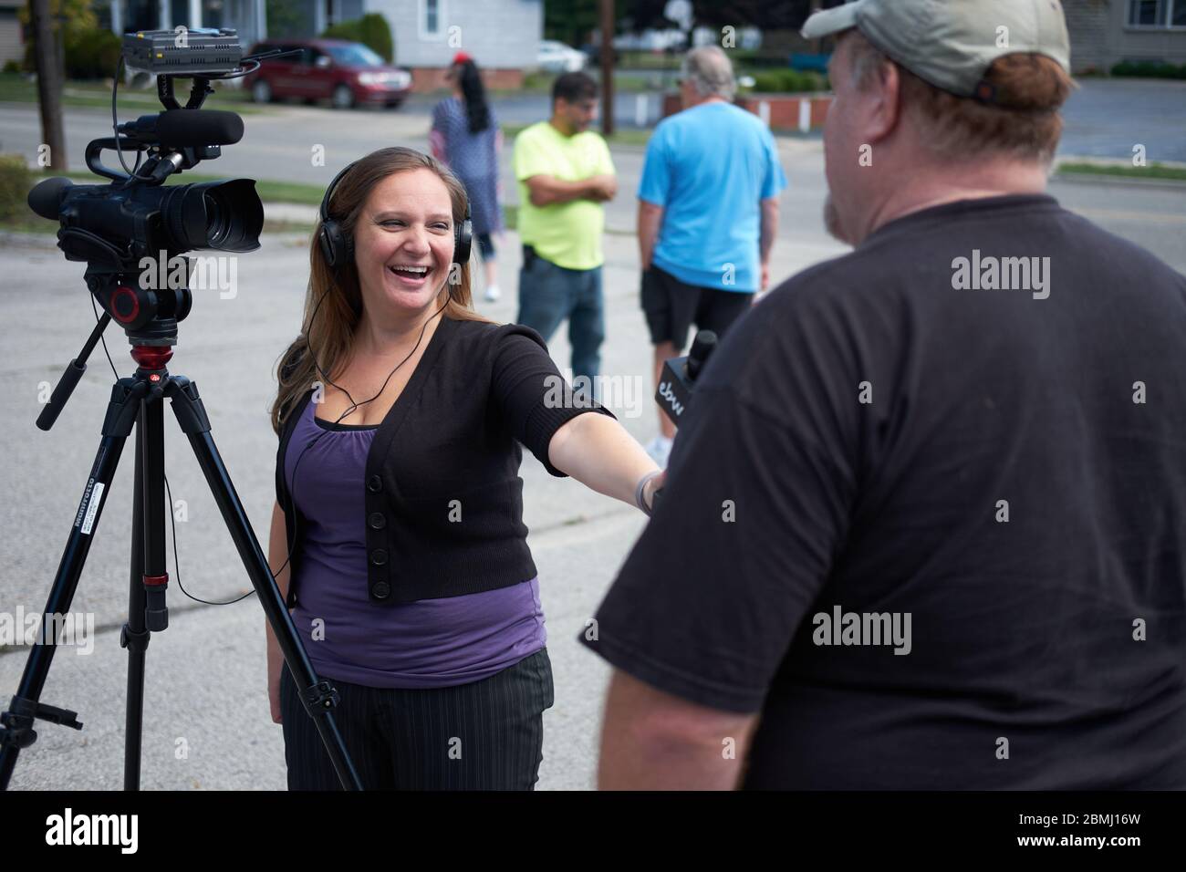 Lone female journalist with great happy smile wearing headphones and holding microphone does an outdoor interview of man in ball-cap wearing t-shirt. Stock Photo