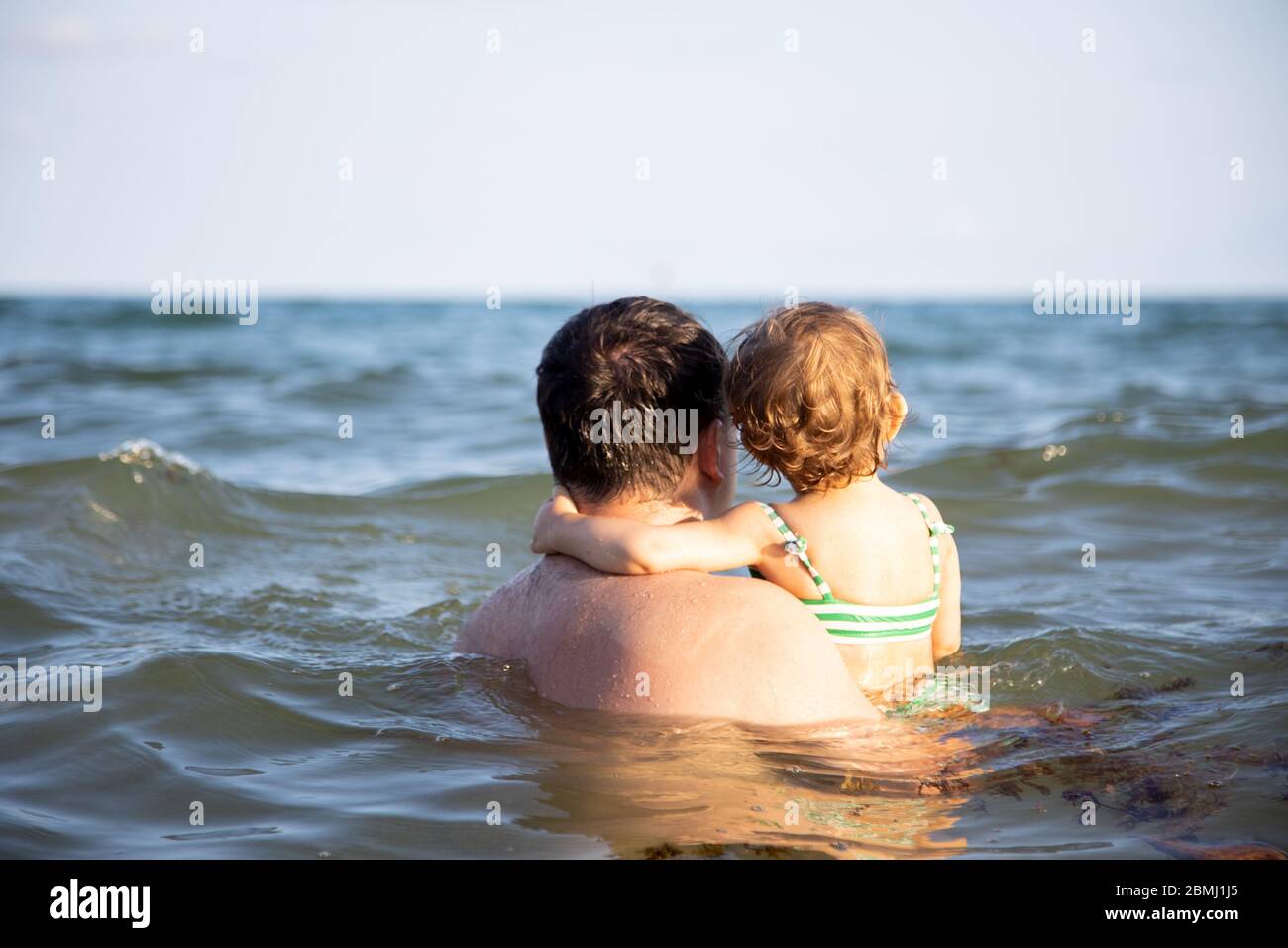 dad holds a little cute toddler daughter in the sea water and watching the horizon back view. Stock Photo