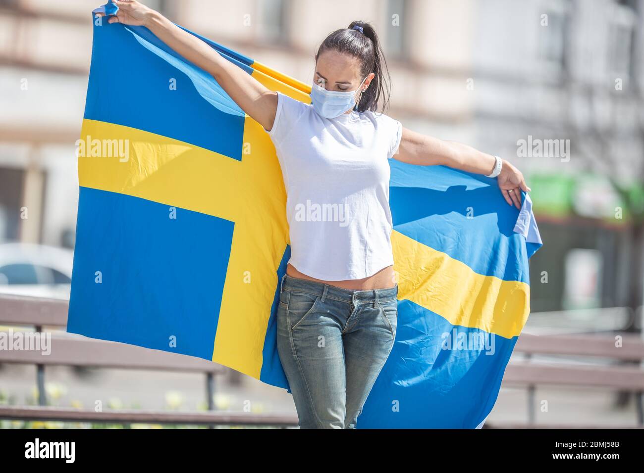 Brunetter standing outdoors looks at the Swedish flag she holds, with a face mask on. Stock Photo