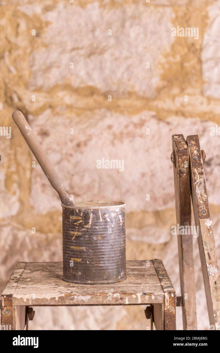 A single paint brush sitting in an old canned fruit tin covered in paint and resting on an old painters ladder in morning sunlight Stock Photo