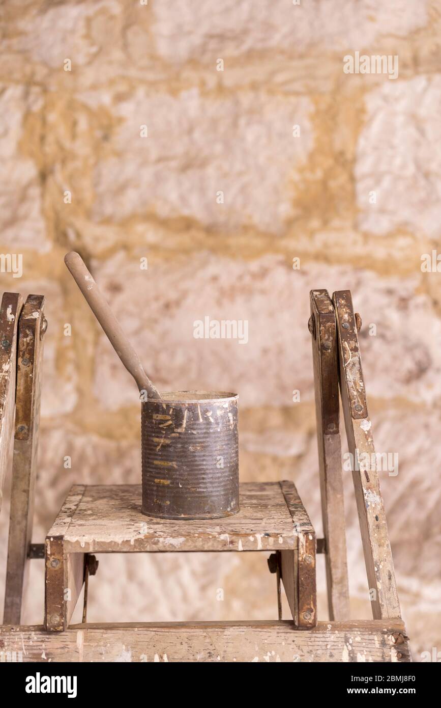 A single paint brush sitting in an old canned fruit tin covered in paint and resting on an old painters ladder in morning sunlight Stock Photo