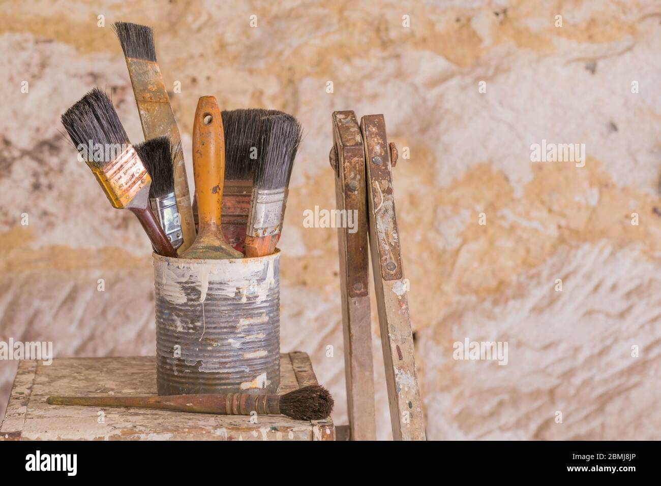 Old paint brushes stacked end up in an old canned fruit tin covered in paint and sitting on an old painters ladder in morning sunlight Stock Photo