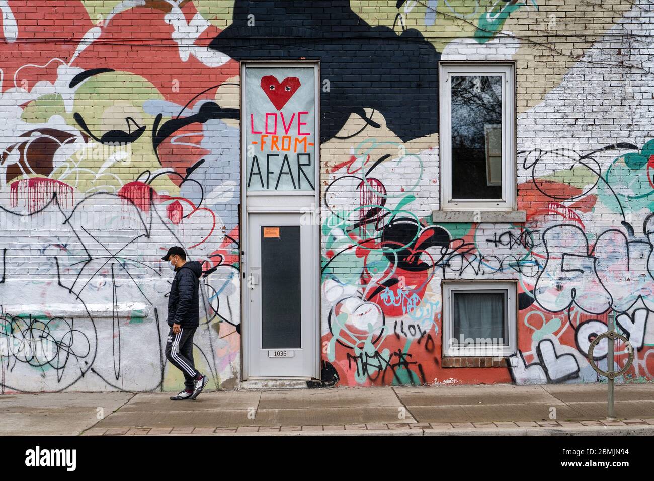 Toronto, Canada, local man wearing face mask walks in front of colourful wall featuring social distancing themed signs during coronavirus pandemic. Stock Photo