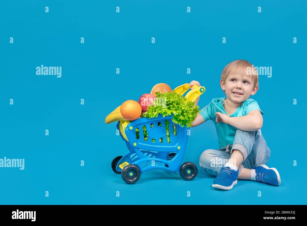 A child of European appearance, a blond boy, is sitting on the floor near a shopping trolley from a supermarket filled with groceries, fruits, bread a Stock Photo