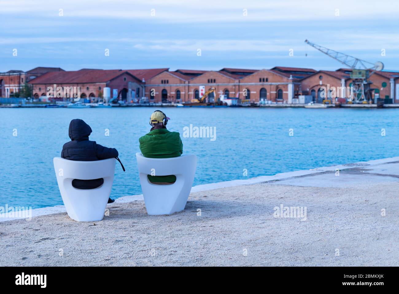 Tourists sitting on white chairs viewing the blurred industrial shipyard with crane along the sea in Venice, Italy Stock Photo