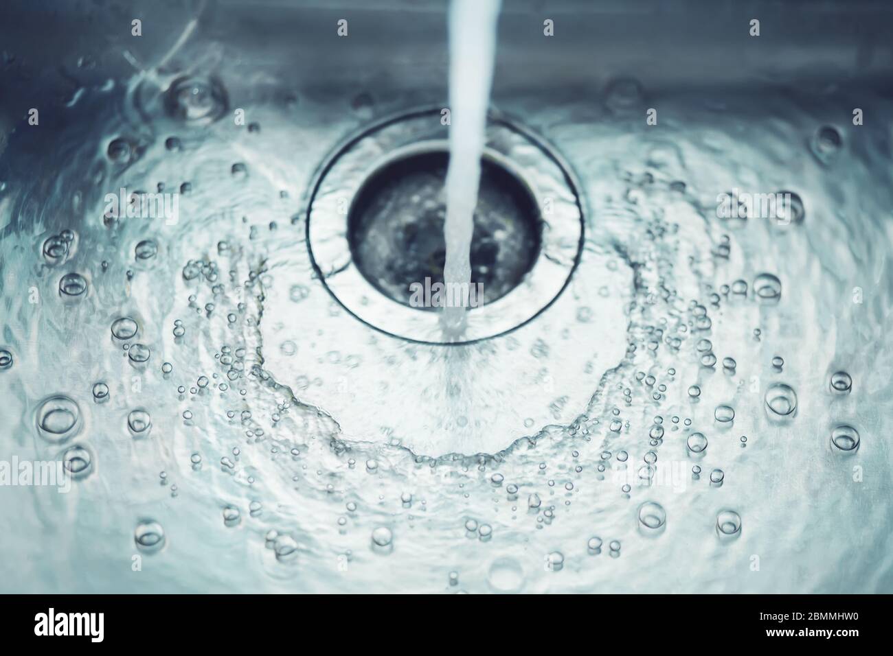 In the metal sink in the kitchen, water flows from the tap, which bubbles and flows into the drain hole. Stock Photo