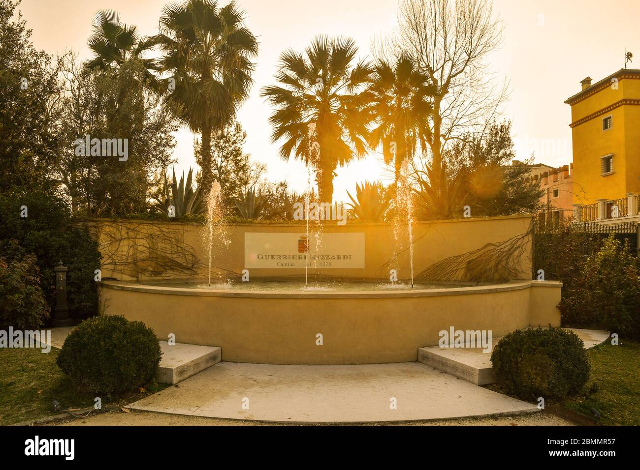 Backlit view of a stone fountain in the park of Villa Guerrieri on the shore of Lake Garda with palm trees at sunset, Bardolino, Verona, Veneto, Italy Stock Photo