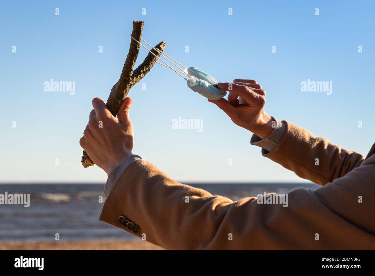Man uses a protective medical mask as a slingshot from tree branch, blue sky on background, selective focus. Enjoys life, back to normal life after co Stock Photo