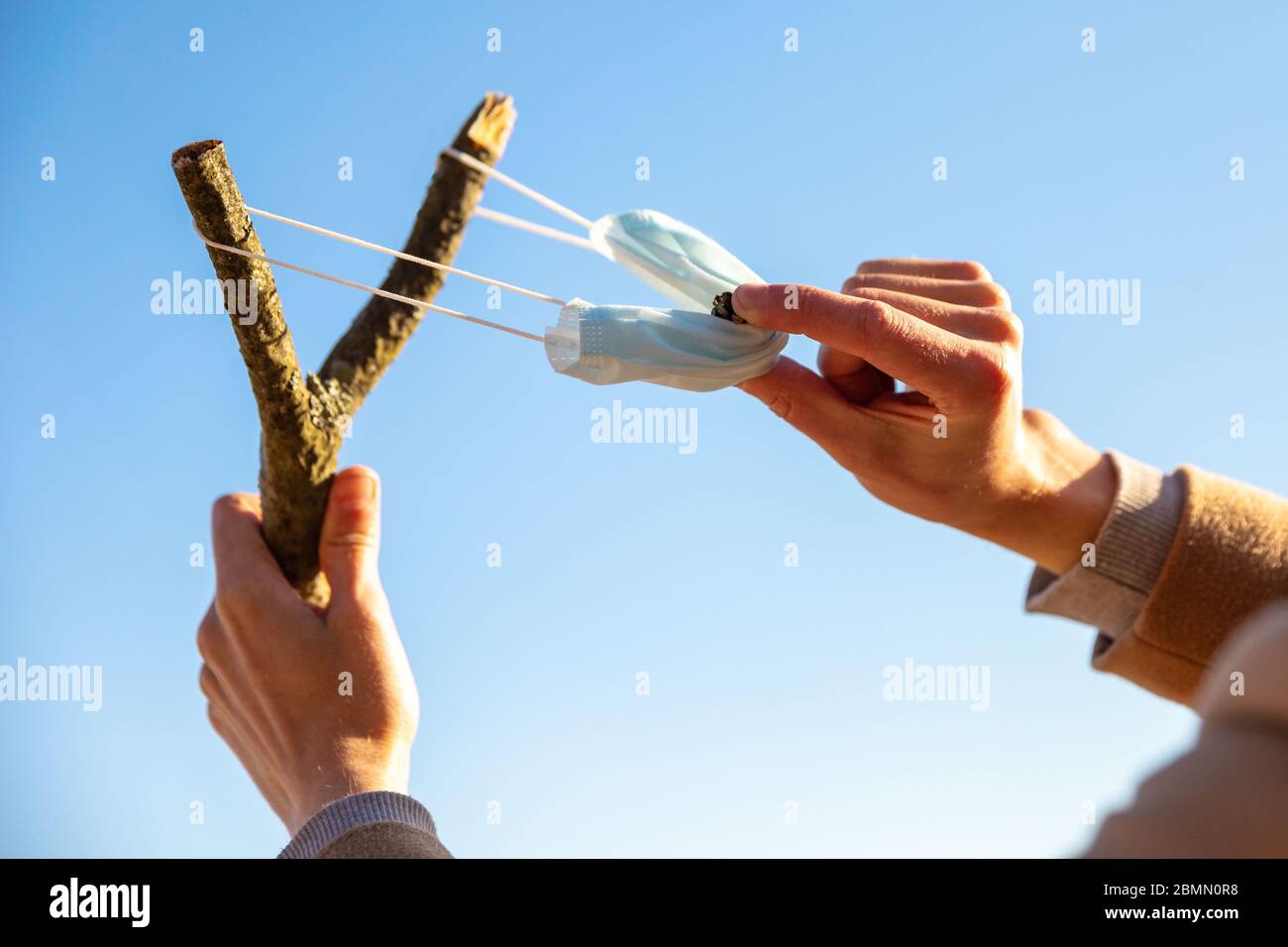 Man uses a protective medical mask as a slingshot from tree branch, blue sky on background, selective focus. Enjoys life, back to normal life after co Stock Photo