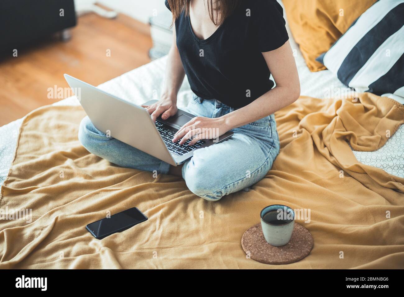 Casual woman working on a laptop sitting on the bed in the house. Female relaxing and drinking cup of hot coffee or tea using laptop computer. Stock Photo